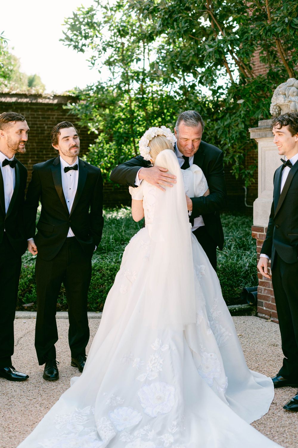 A wedding party shares an emotional moment outdoors with the bride in a flowing white gown.