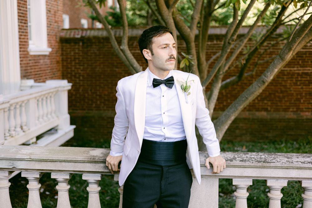 A person in a white tuxedo jacket and bow tie poses against a brick wall with greenery.
