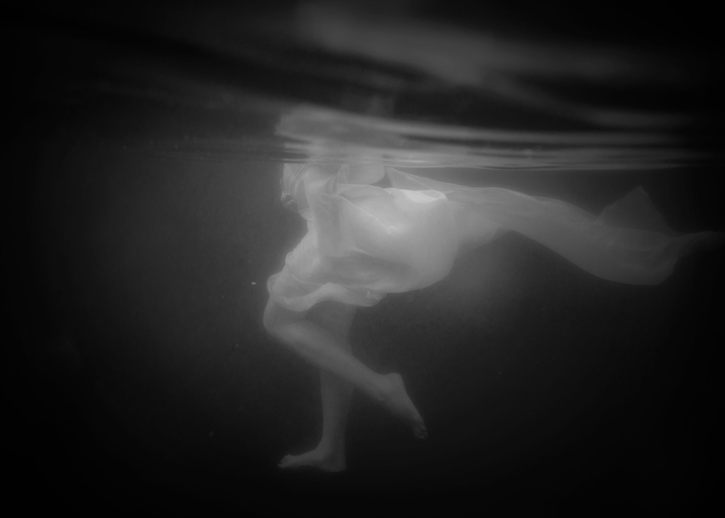 Black and white underwater photoshoot of lady walking with white dressing floating behind.