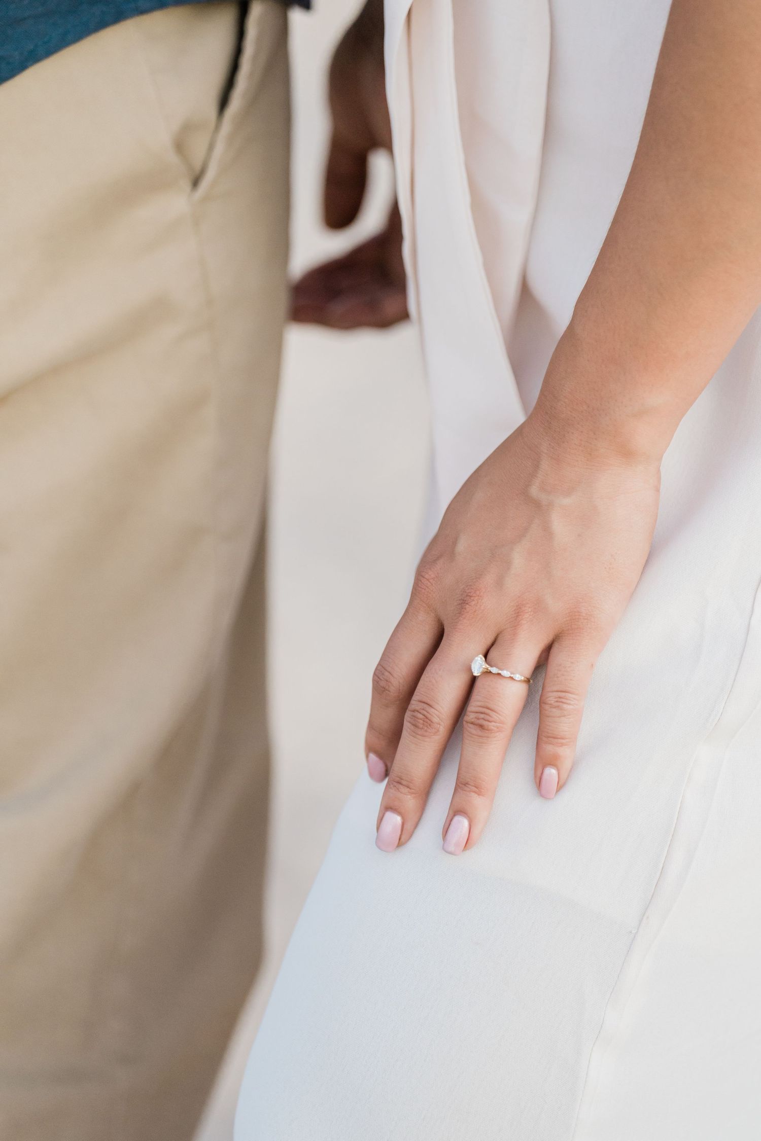 Close-up of a bride's hand showcasing an elegant engagement ring against a white dress.