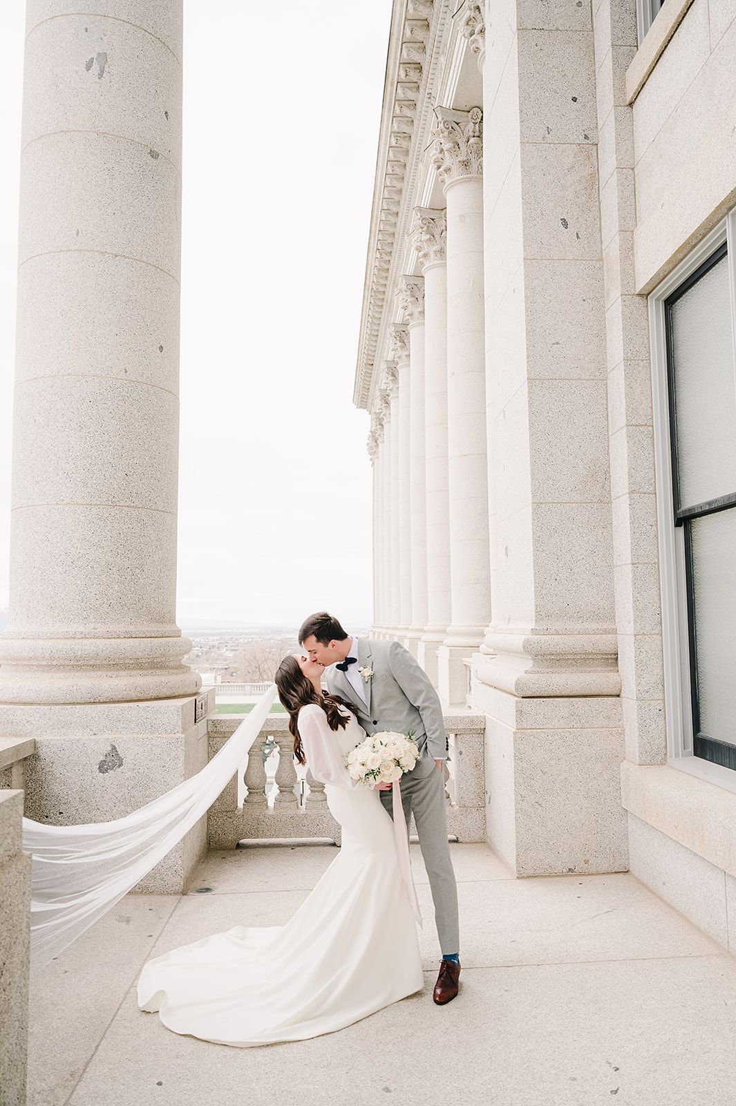 Romantic wedding pose with flowing veil between classical white columns of historic building.