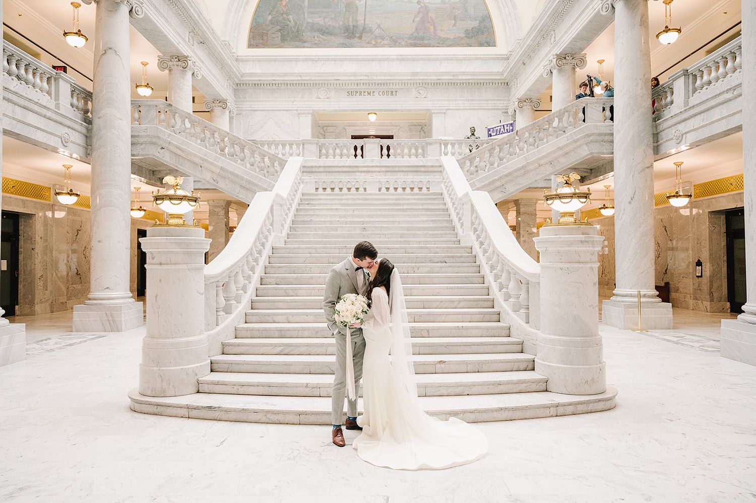 Wedding couple embracing on grand white marble staircase in elegant historic building interior.