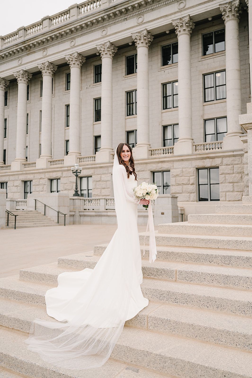 Bride in flowing white gown poses on elegant stone steps of historic building.