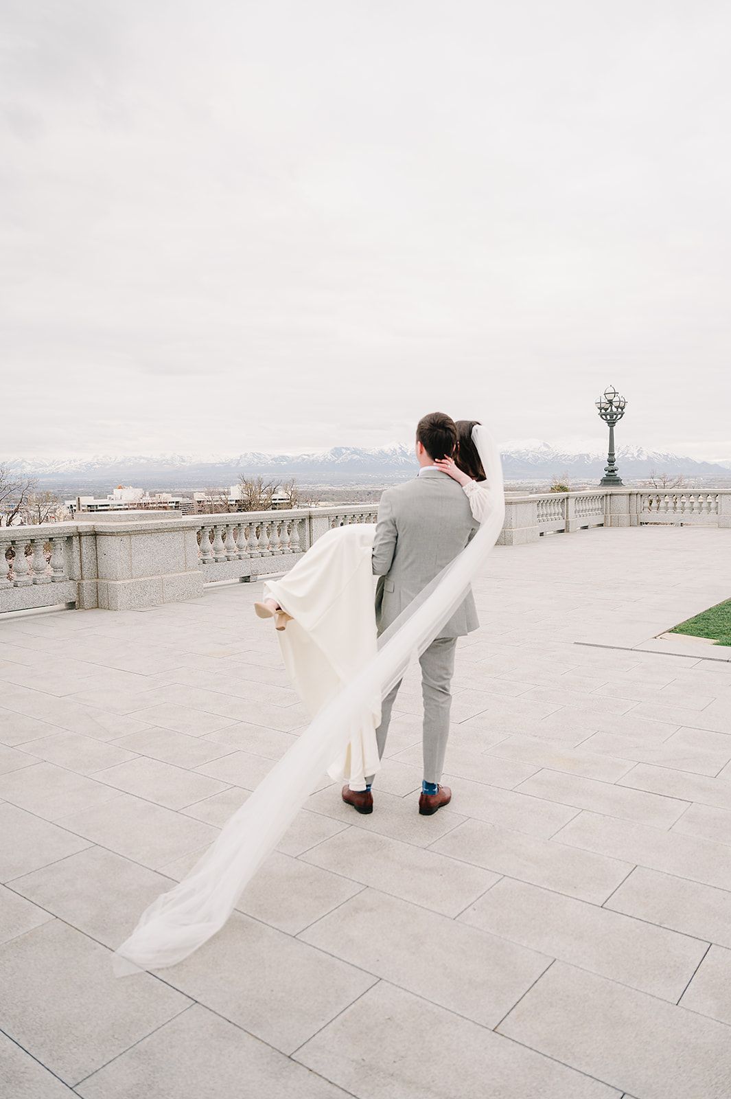 Couple in wedding attire stands on balcony overlooking mountain landscape.