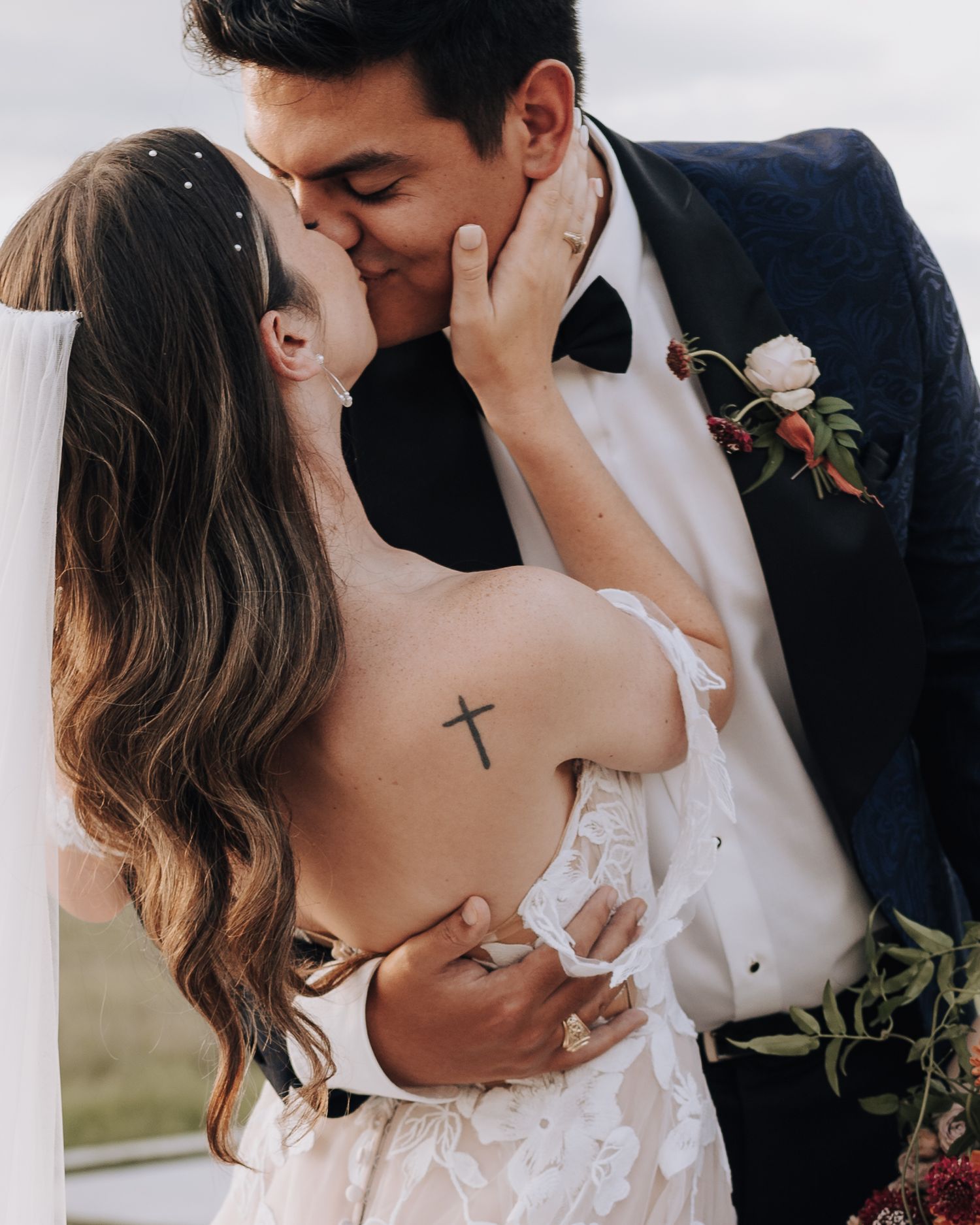 A romantic wedding day kiss captured with a cross tattoo visible on the bride's back.