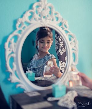Elegant white baroque mirror on turquoise wall reflects vanity scene with jewelry and decorative items.
