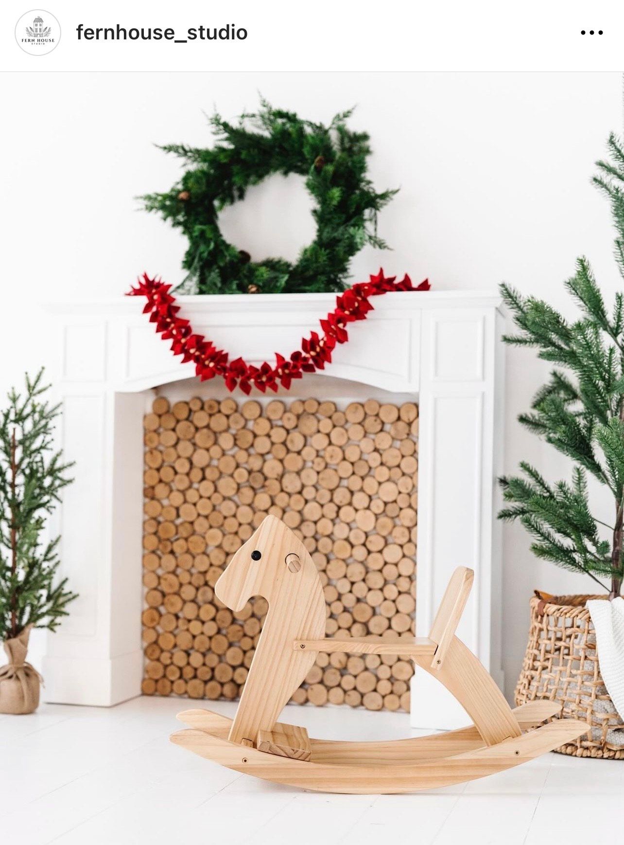 Wooden rocking horse beside a decorative fireplace with stacked logs and Christmas wreath.