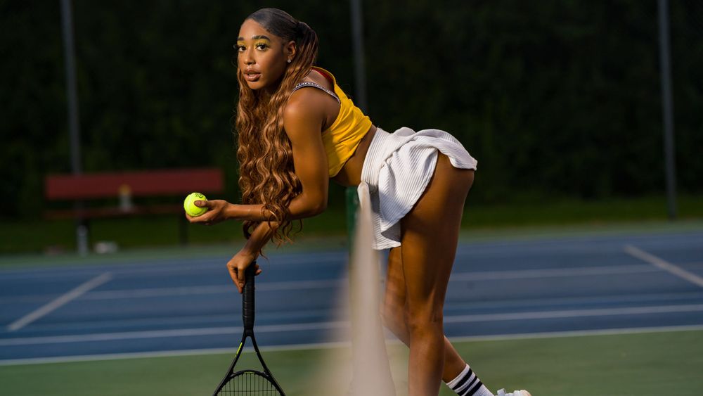 Model in yellow crop top and with skirt on tennis court with racket leaning on the net with a ball in hand photoshoot