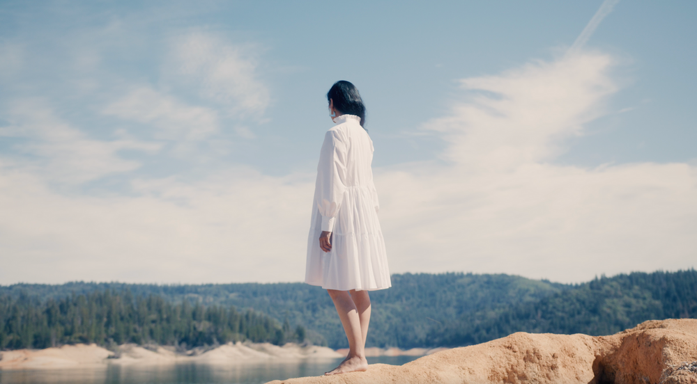 Mysterious woman in white dress standing mountain high overlook a forest of trees and blue waters in video production