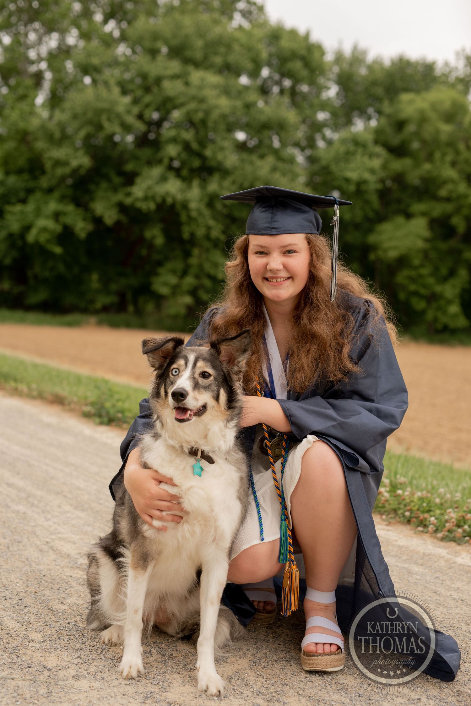 Graduate in blue cap and gown sits on dirt path with their husky mix companion.