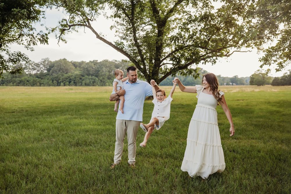Family of four playing in a green field under a large tree, parents swinging young child while holding toddler.