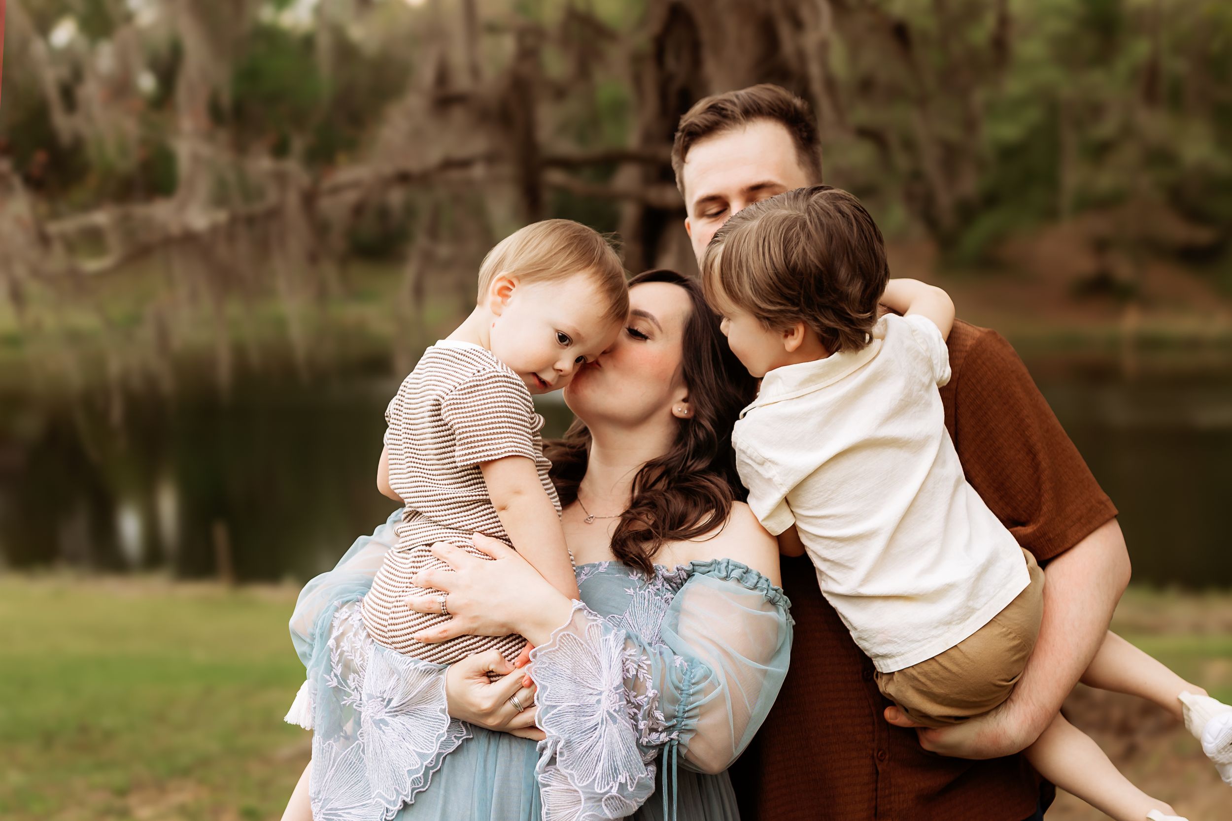 Family portrait of parents kissing their two young children outdoors near a pond surrounded by trees.