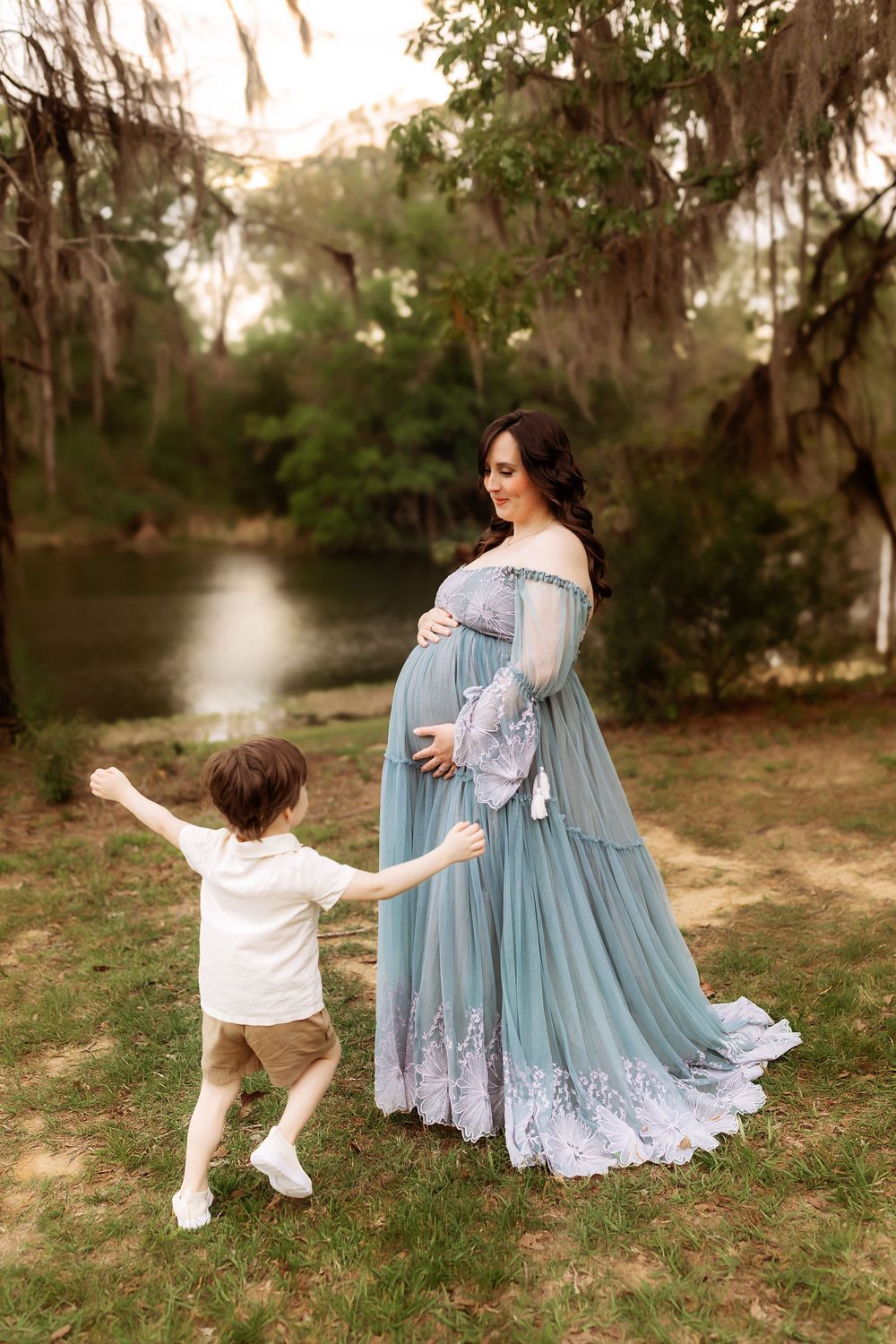 Pregnant woman in teal gown holds hands with toddler boy outdoors near a serene river surrounded by lush trees.