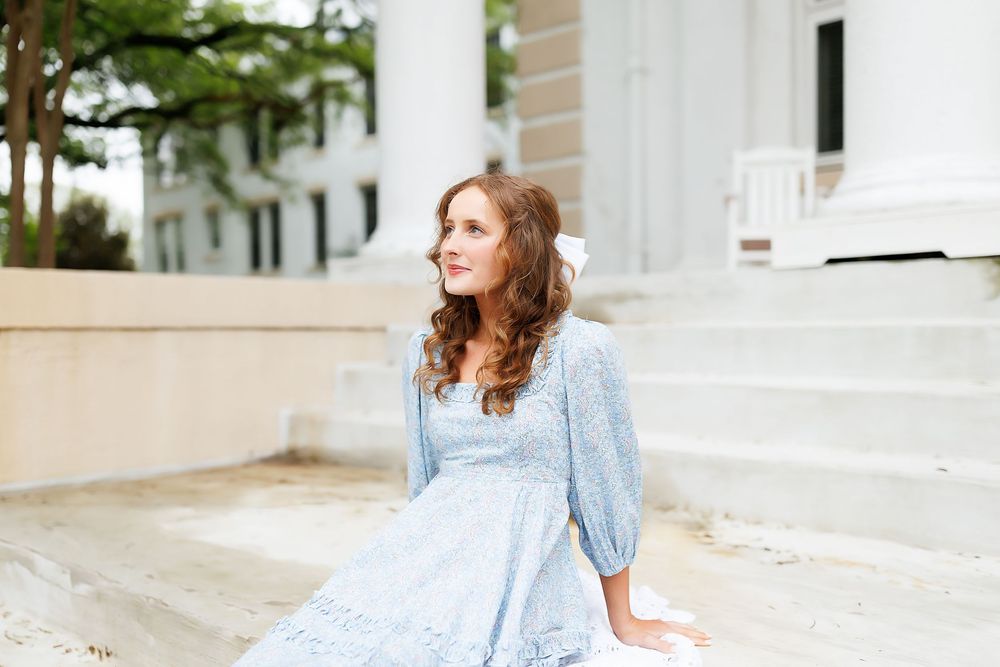 A serene figure in a light blue vintage dress sits on stone steps outside a classic white columned building.