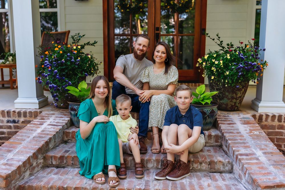 A family sits together on brick front porch steps surrounded by colorful flower planters.
