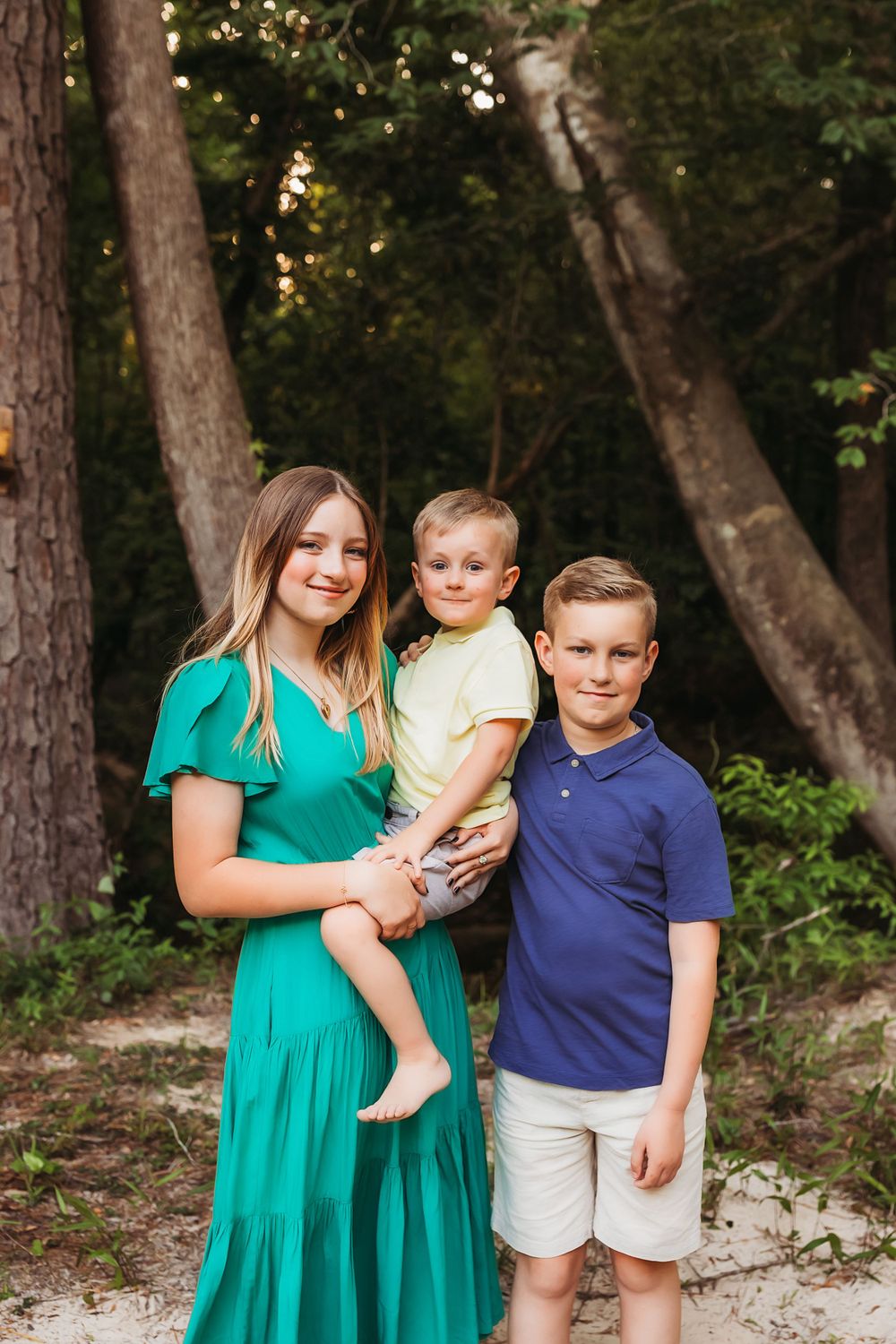Three siblings pose together outdoors with trees in the background at sunset.