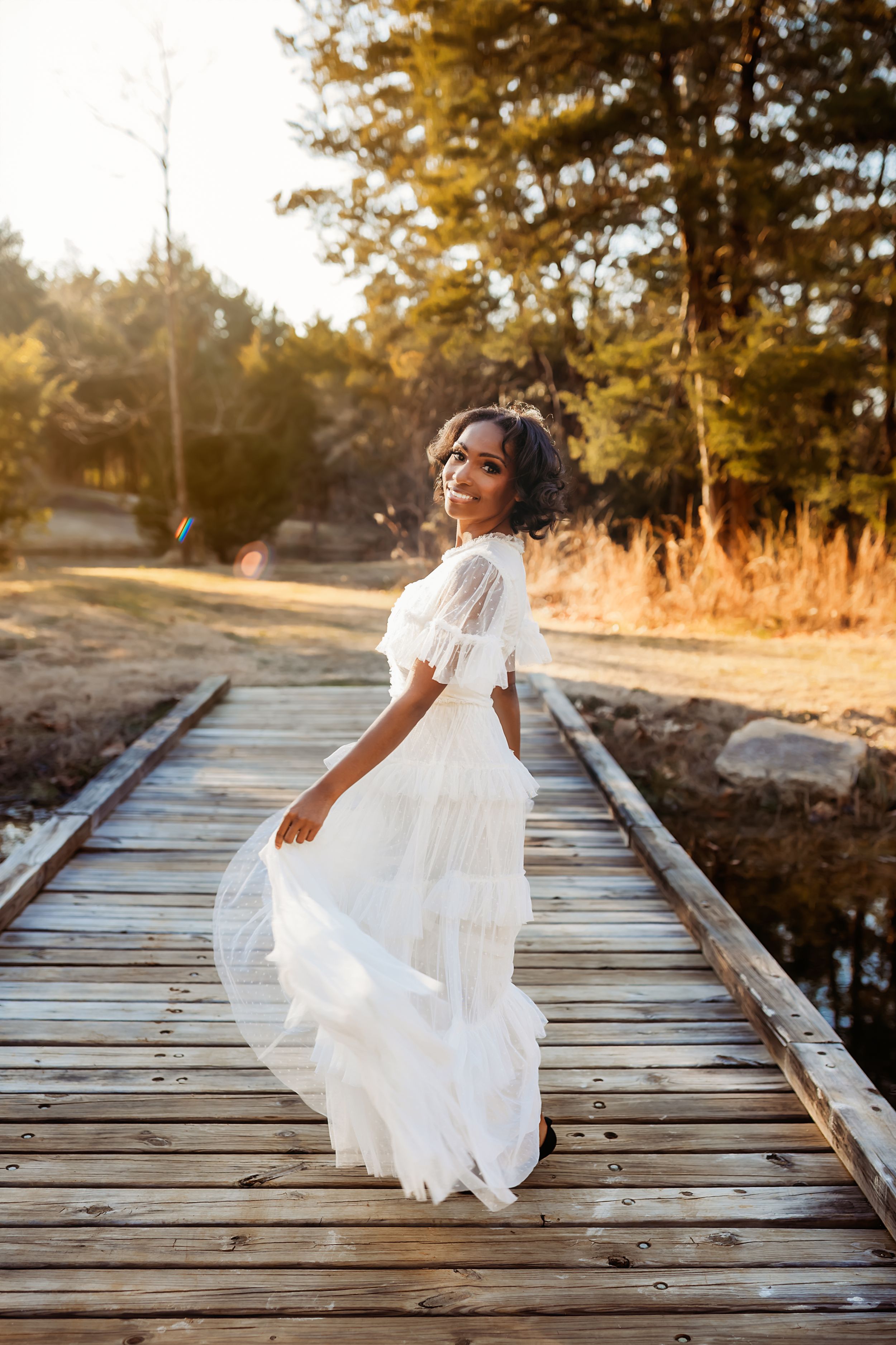 A flowing white dress catches the sunlight on a wooden bridge surrounded by golden autumn trees at sunset.