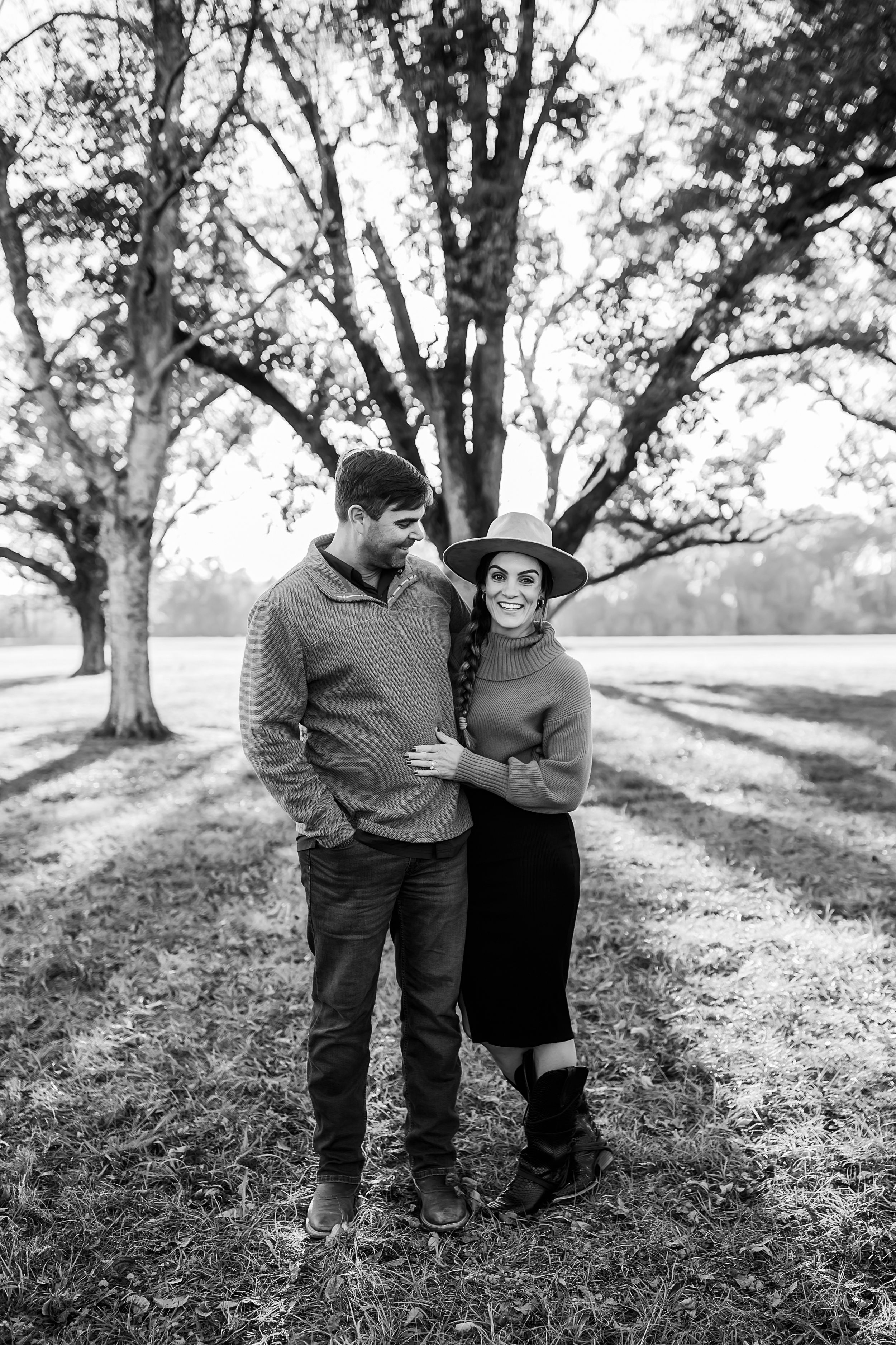 A couple shares an intimate moment in a park with tall trees in black and white.
