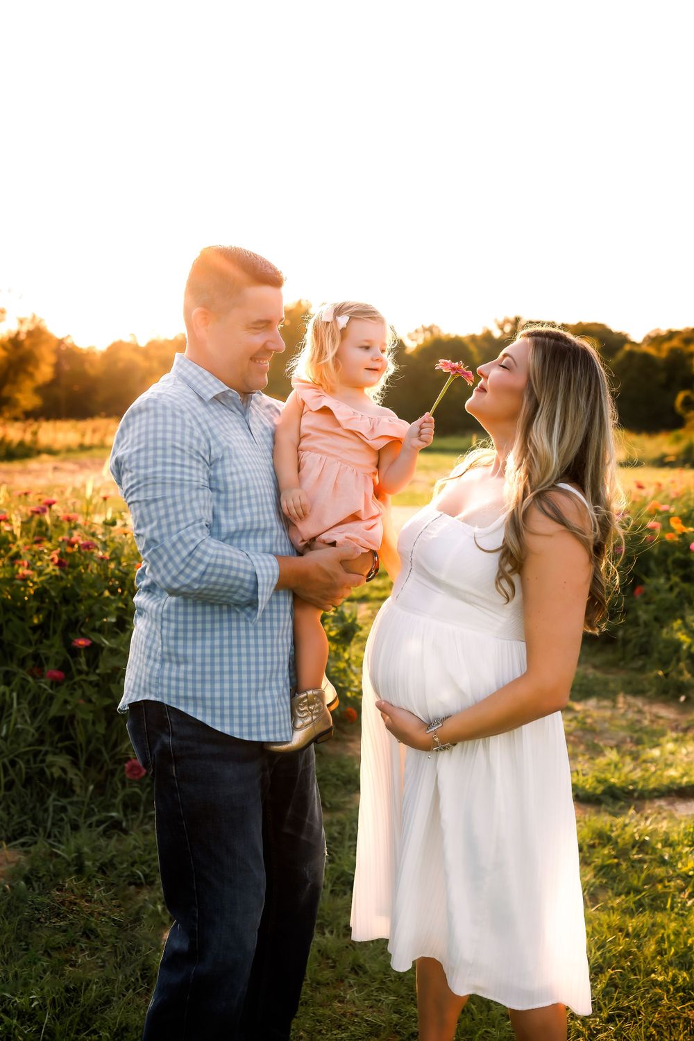 Family shares joyful moment at sunset in field with pregnant mother in white dress.