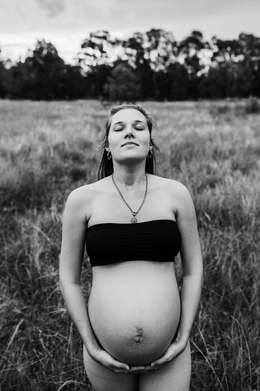 Black and white maternity portrait in field with trees silhouetted against sky.