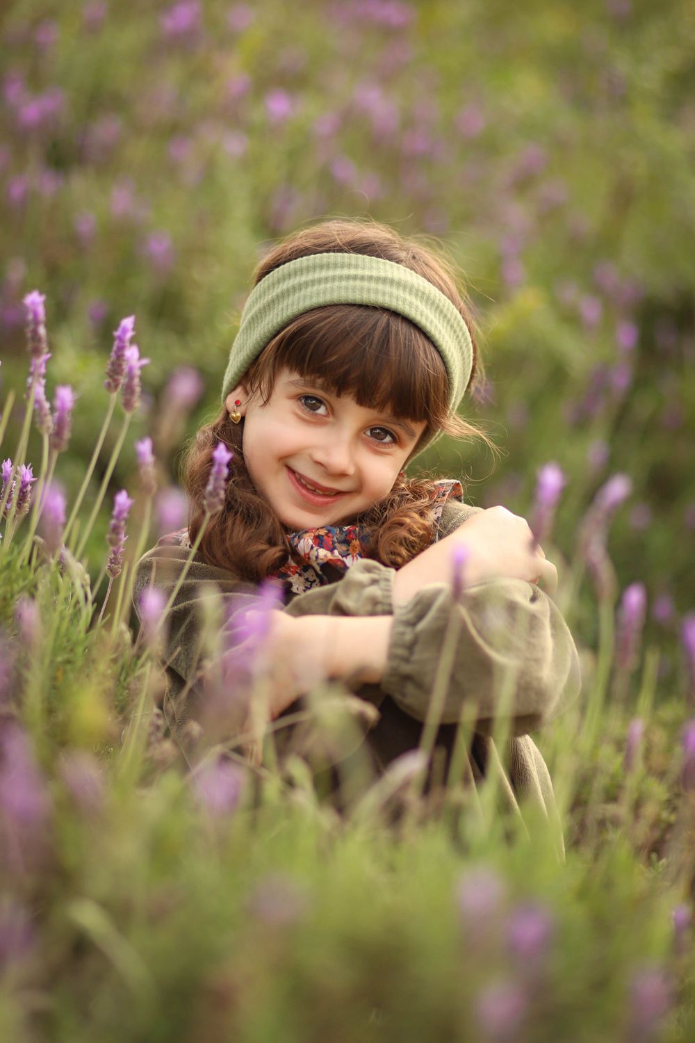 Child wearing a headband sits among purple lavender flowers in a natural outdoor setting.