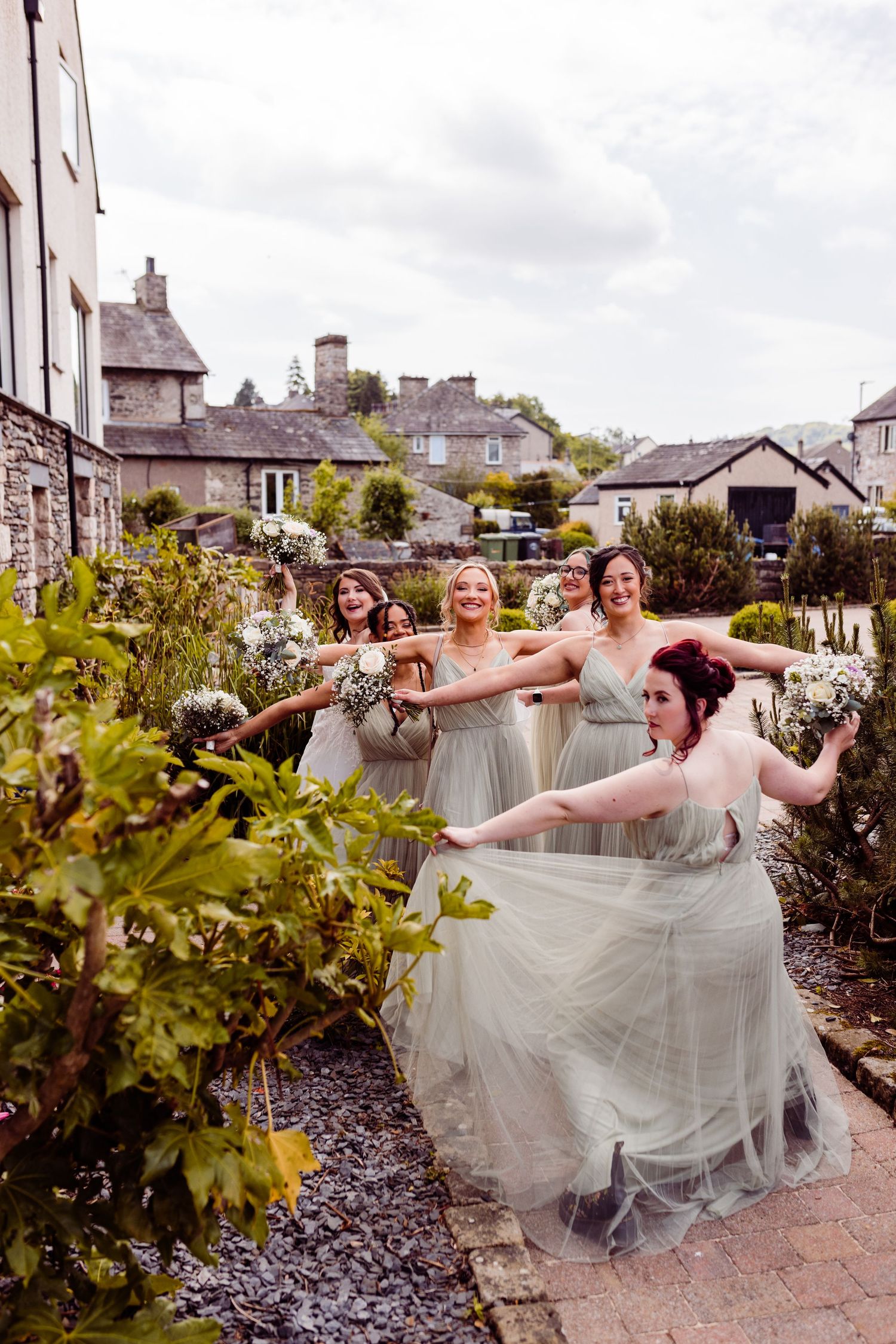 Bridesmaids in light gray dresses pose playfully in garden setting.