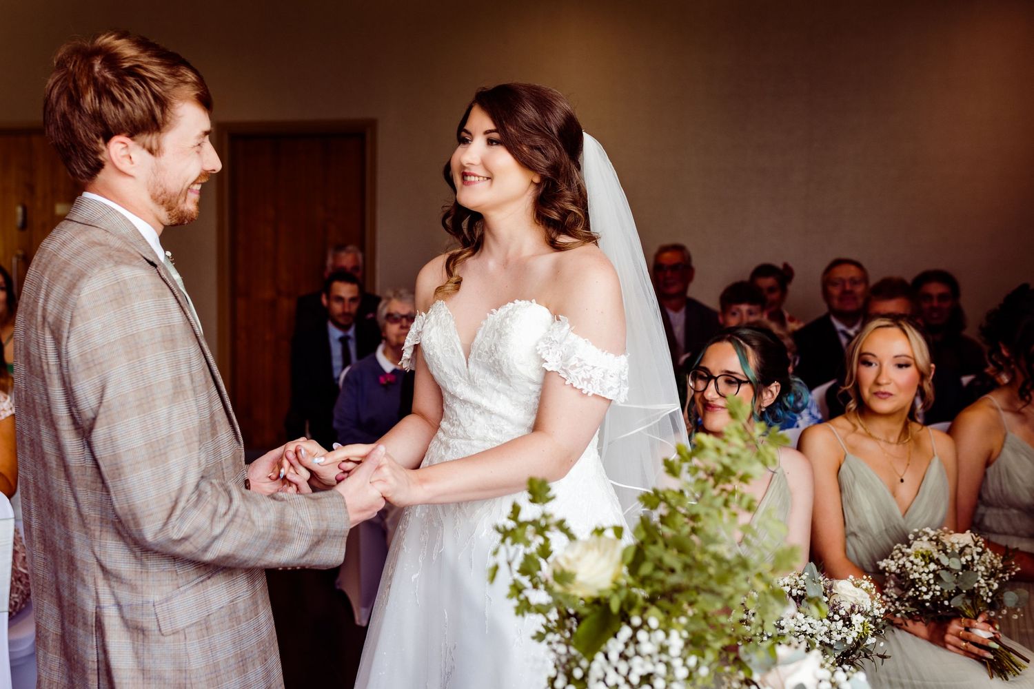 A couple exchanges rings during their wedding ceremony surrounded by guests and floral decorations.