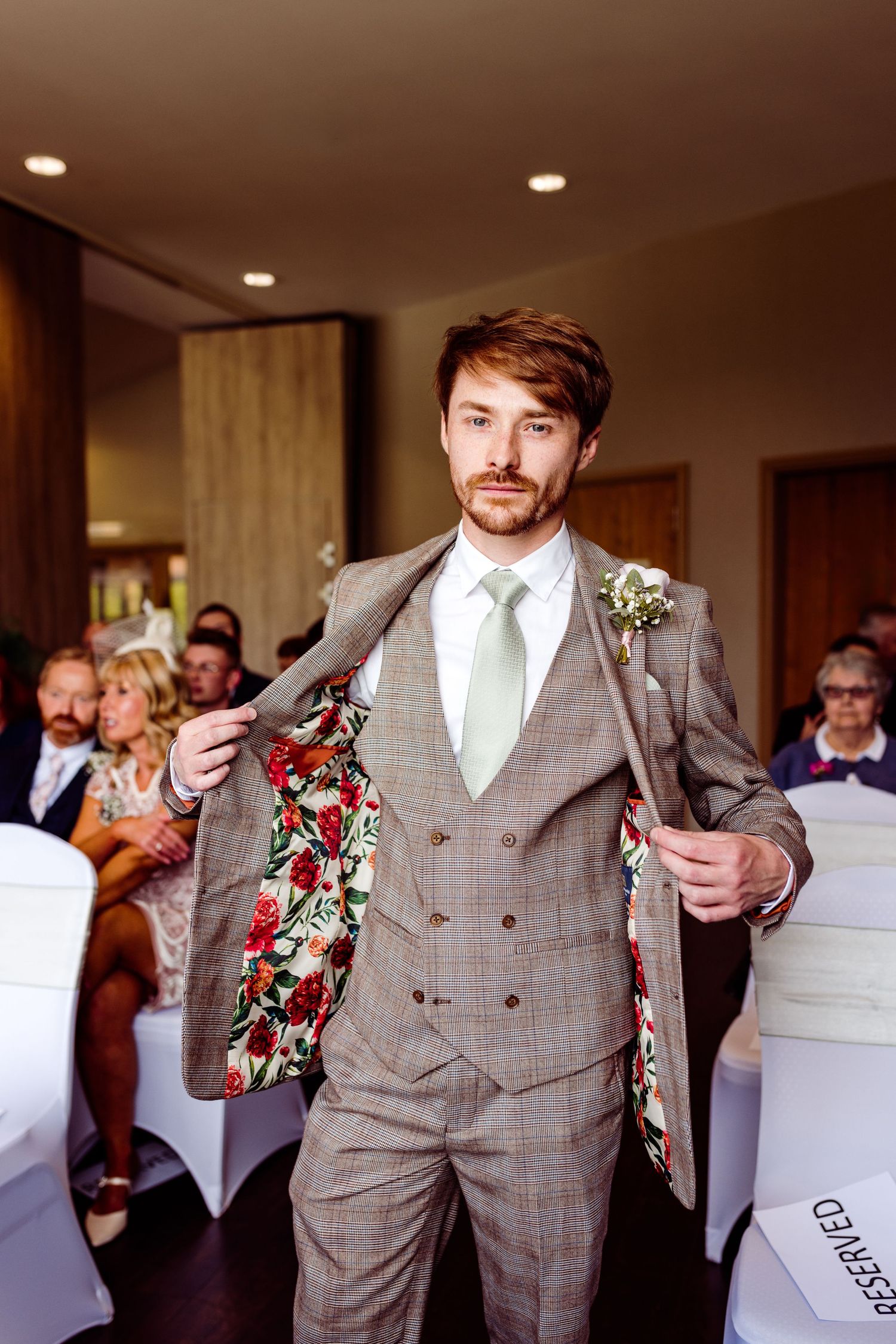 Groom in a stylish gray plaid suit reveals colorful floral lining at an indoor wedding venue.