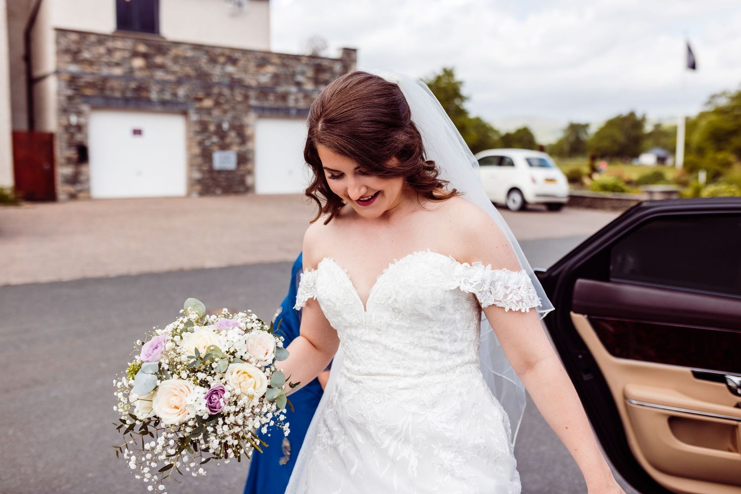 Wedding attendee in off-shoulder white dress holding floral bouquet outside stone building.