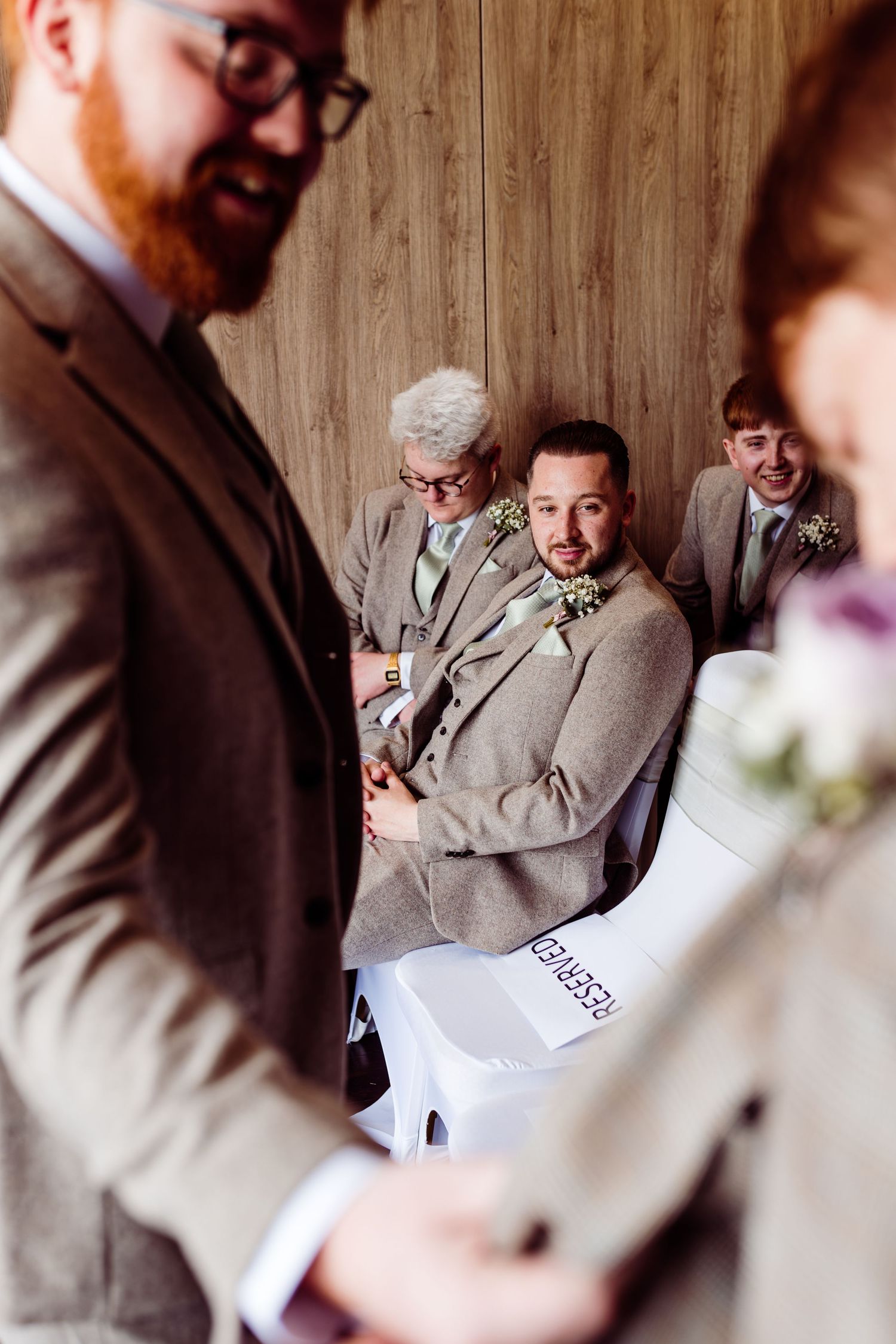 Wedding guests in gray suits gather during a ceremony as they look at papers or programs.