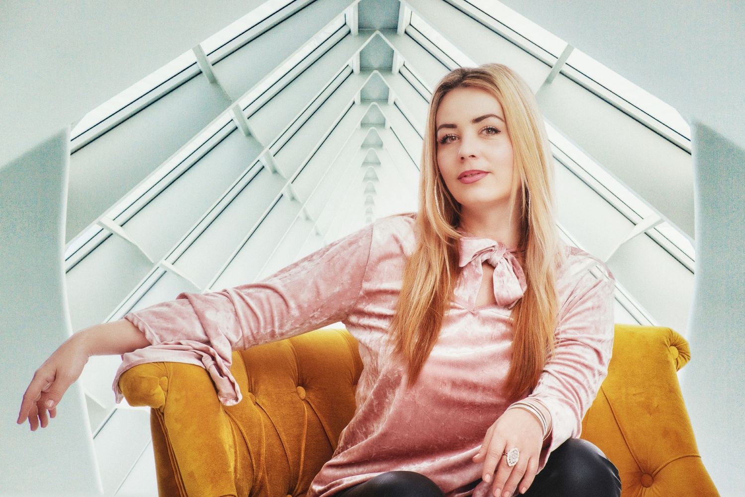 Portrait of a person in pink sitting on a yellow chair under modern skylight architecture.