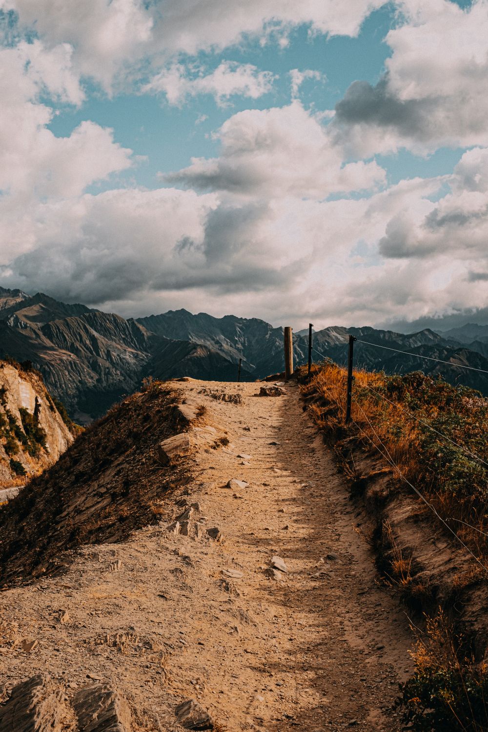 A dirt hiking trail winds along a mountain ridge with wooden fence posts under dramatic cloudy skies.