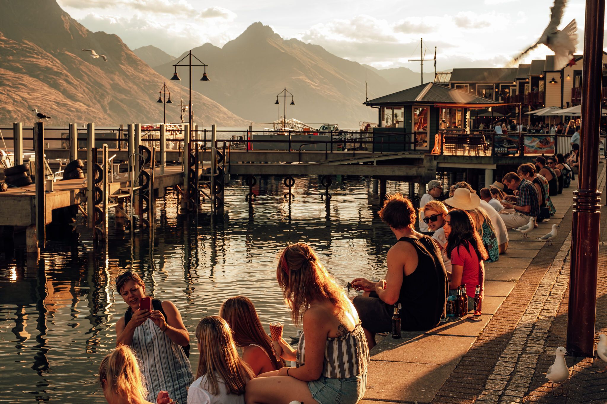 Tourists relax along a scenic waterfront pier with dramatic mountain peaks and warm sunset lighting in Queenstown, New Zealand.