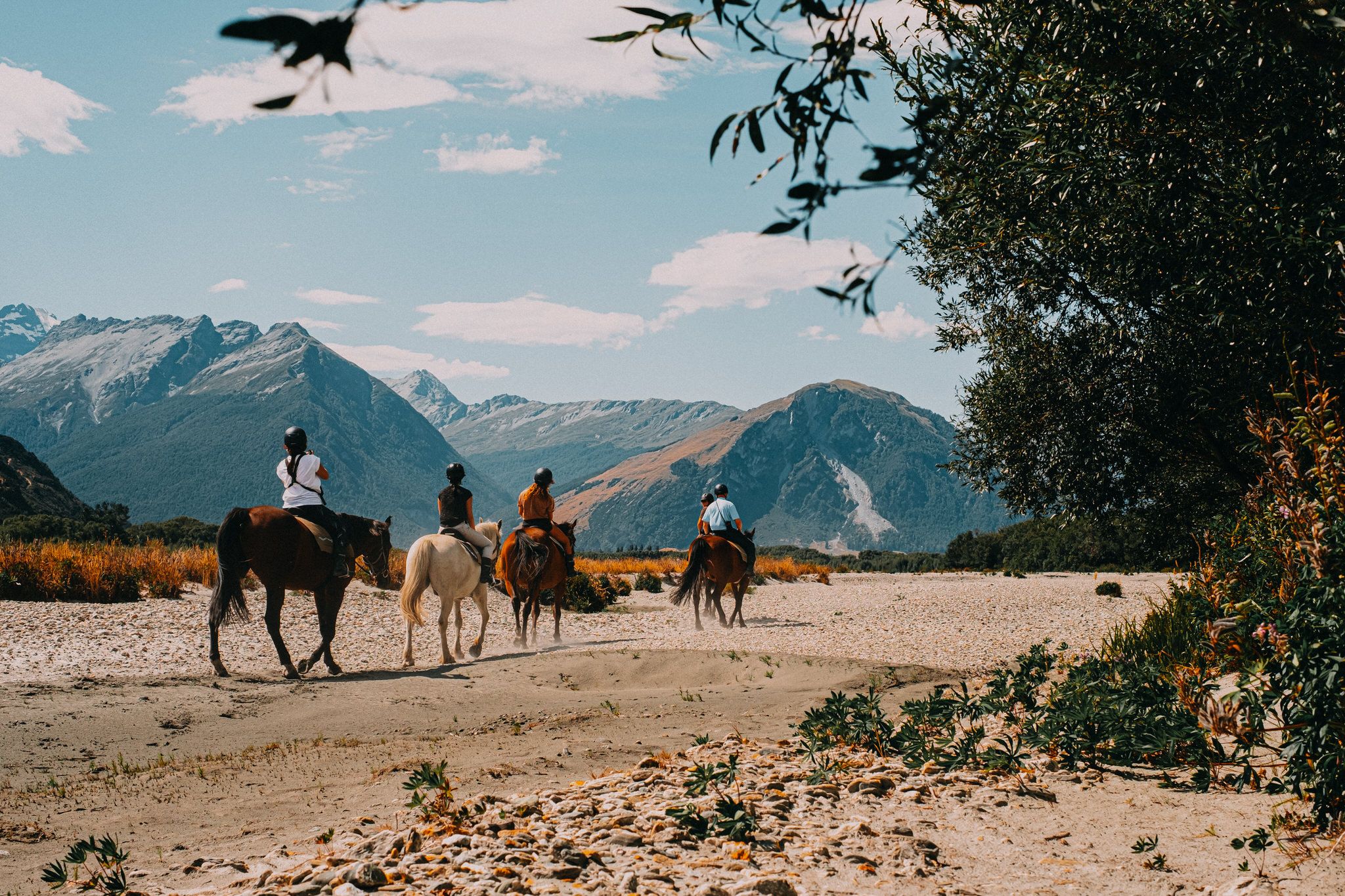 A group of riders on horseback travels along a dirt path with majestic mountains and autumn foliage in the background.