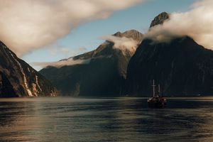 A vintage sailboat cruises through Milford Sound in New Zealand with dramatic misty mountains rising from the water.