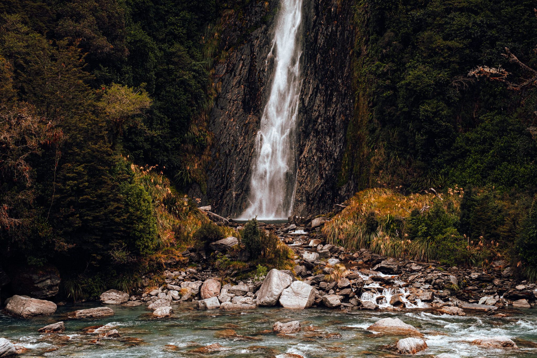 A tall waterfall cascades down a rocky cliff into a mountain stream surrounded by autumn foliage in New Zealand.