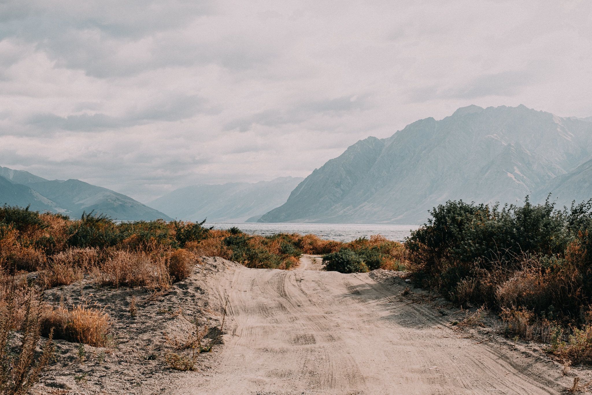 A dusty dirt road winding through arid desert landscape with dramatic mountains and cloudy skies in the background.