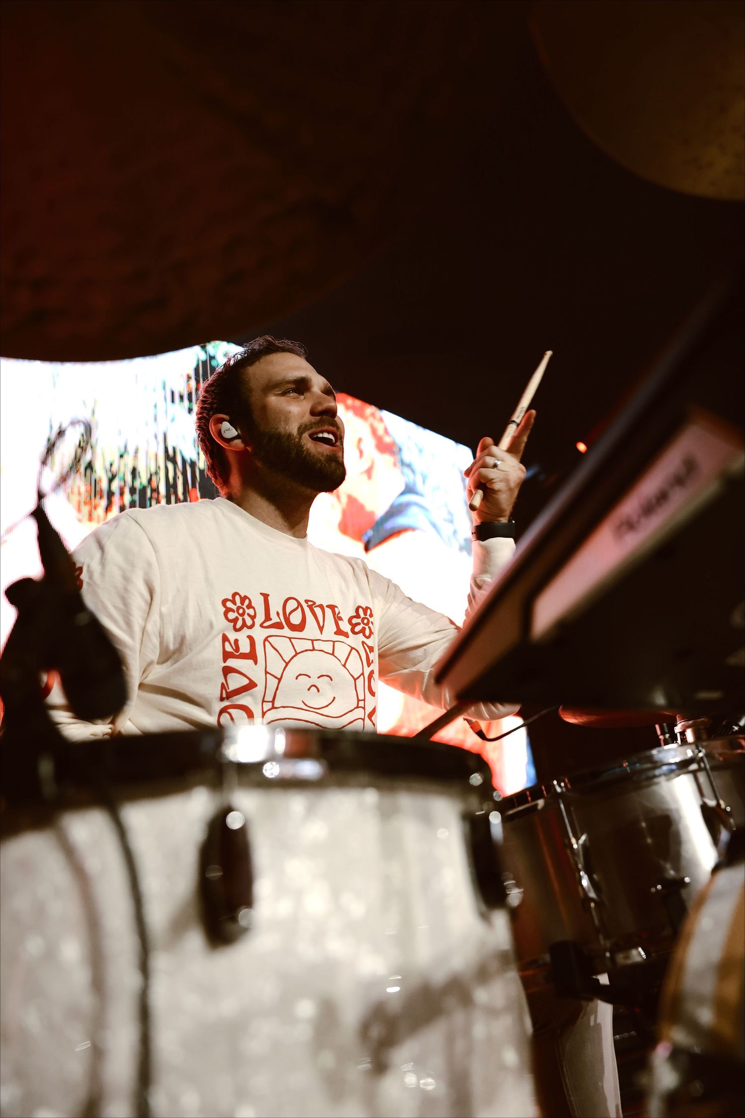An energetic drummer performing on stage wearing a white graphic t-shirt.