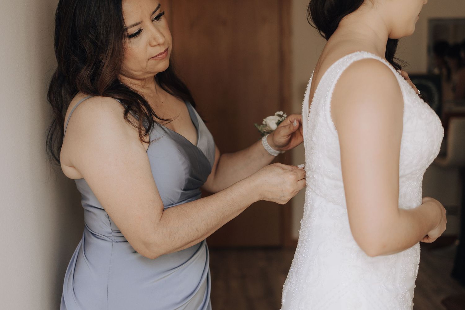 A bridesmaid in gray helps adjust the back of a white wedding dress during preparation.