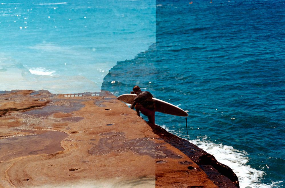 Person lying on rocky cliff edge overlooking deep blue ocean waters.