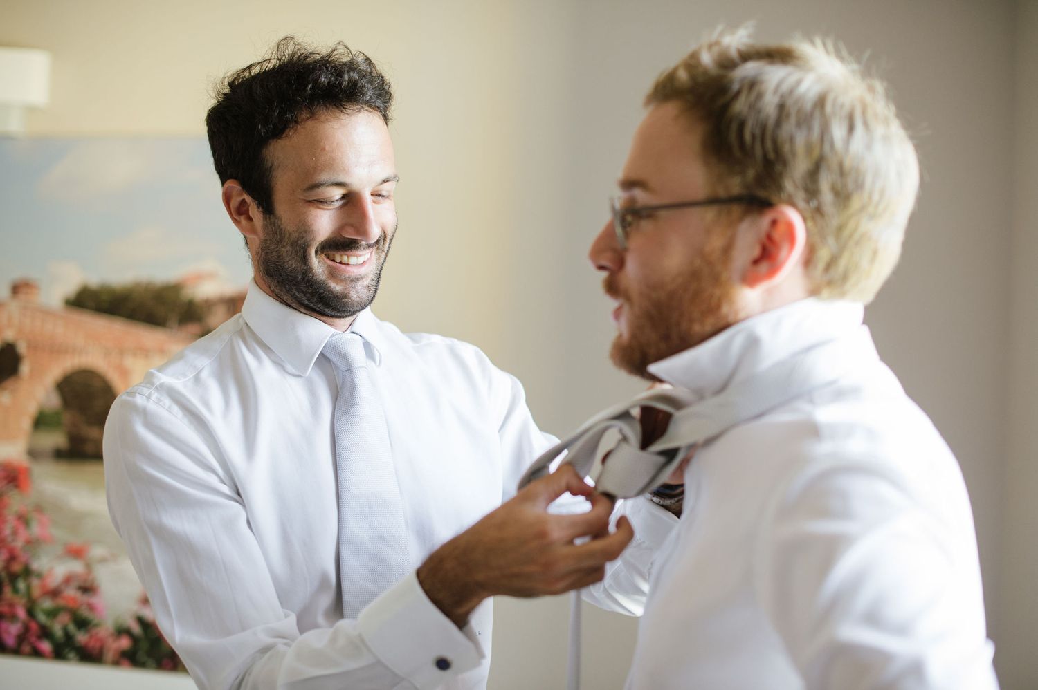 Two people adjusting formal wear and laughing together in a bright room.