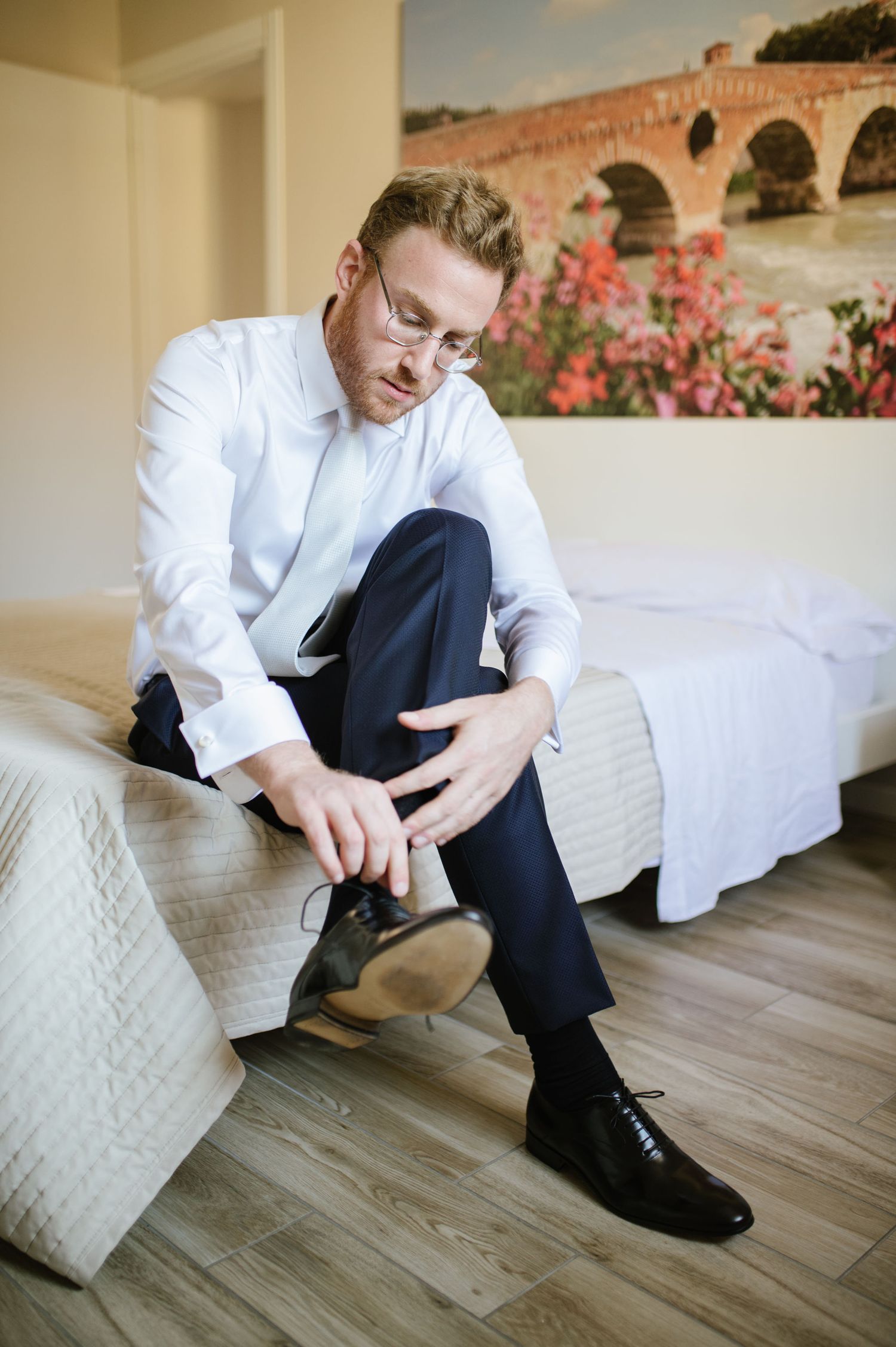 Person in formal white shirt sitting on bed while putting on dress shoes.