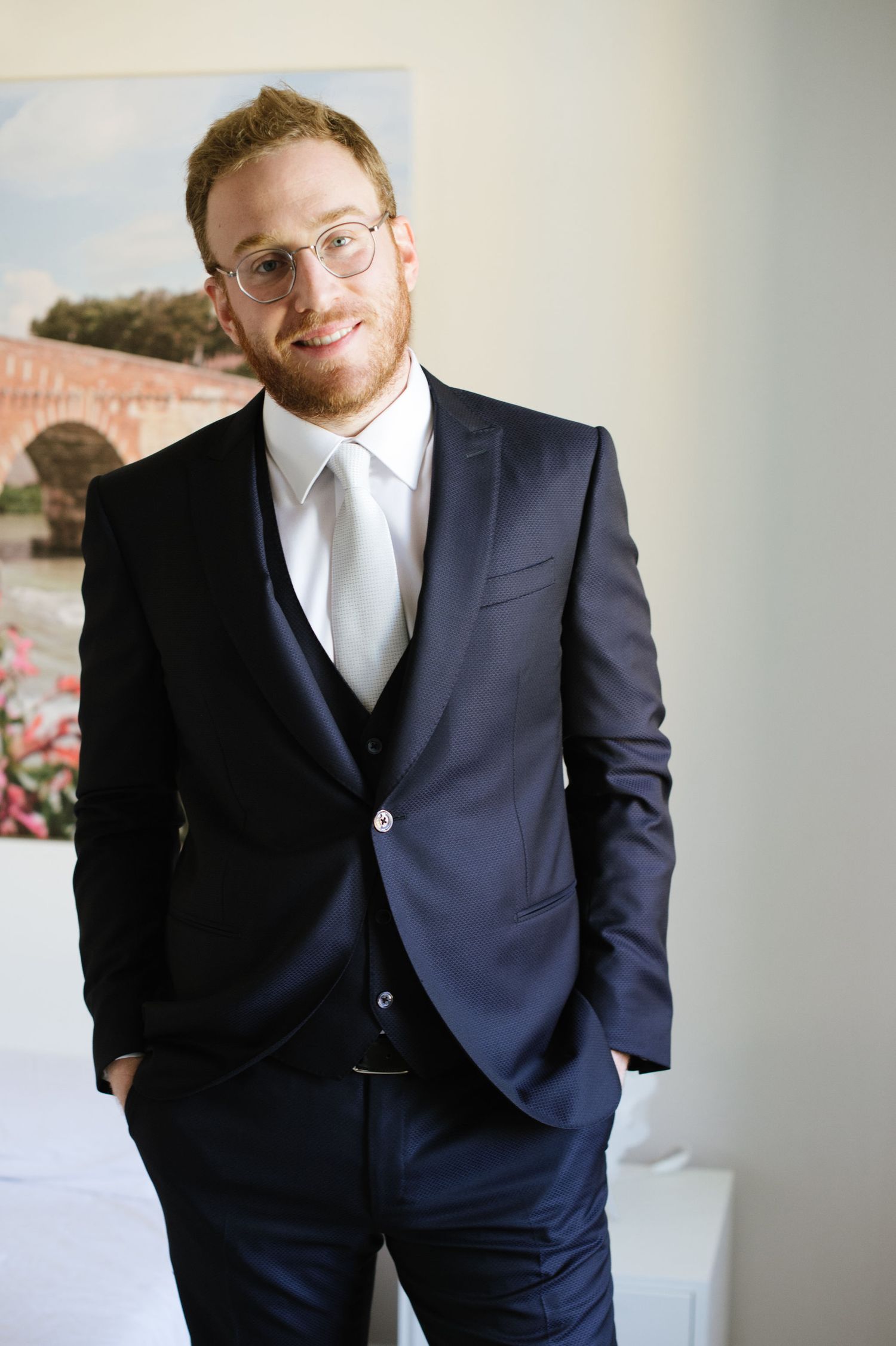 Person wearing navy suit and tie standing against wall with bridge artwork.