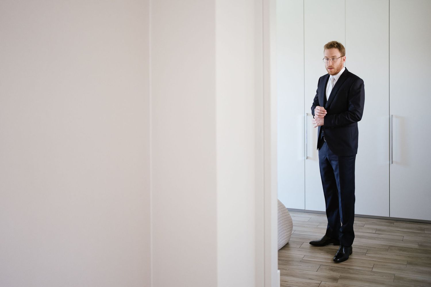 Person in dark formal suit standing in minimalist white room corner.