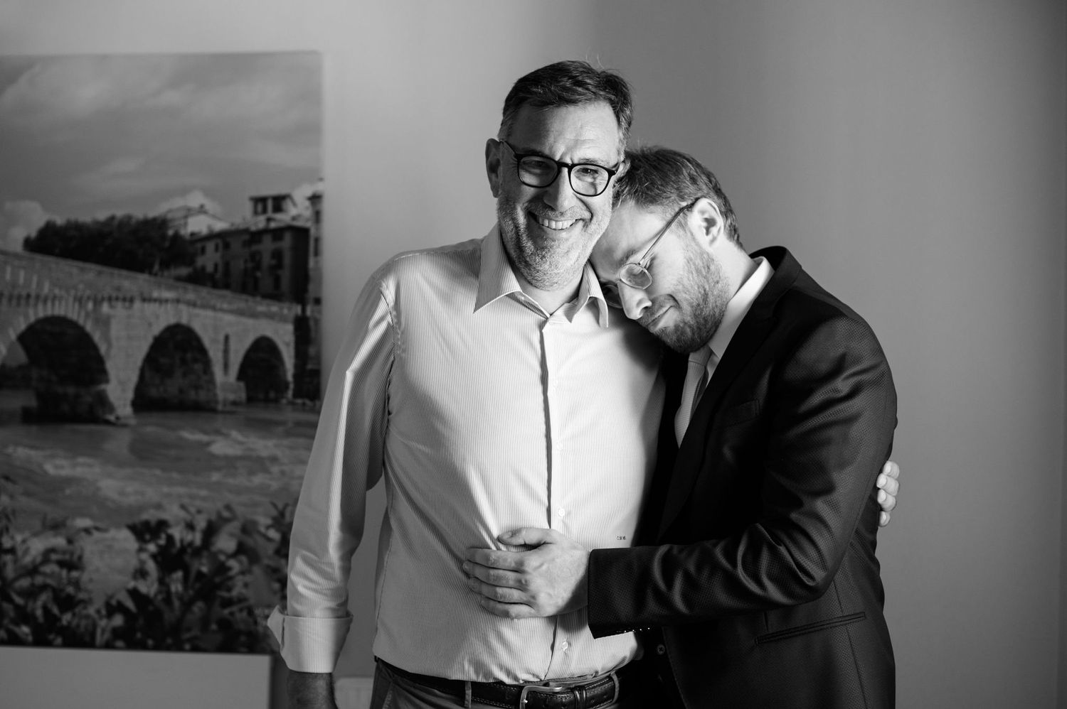 Two people share an intimate black and white moment in front of a framed landscape artwork with an arched bridge.