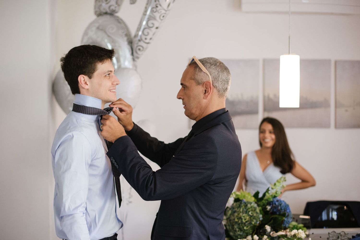 A person in navy suit adjusts another's tie while someone holds flowers in the background.