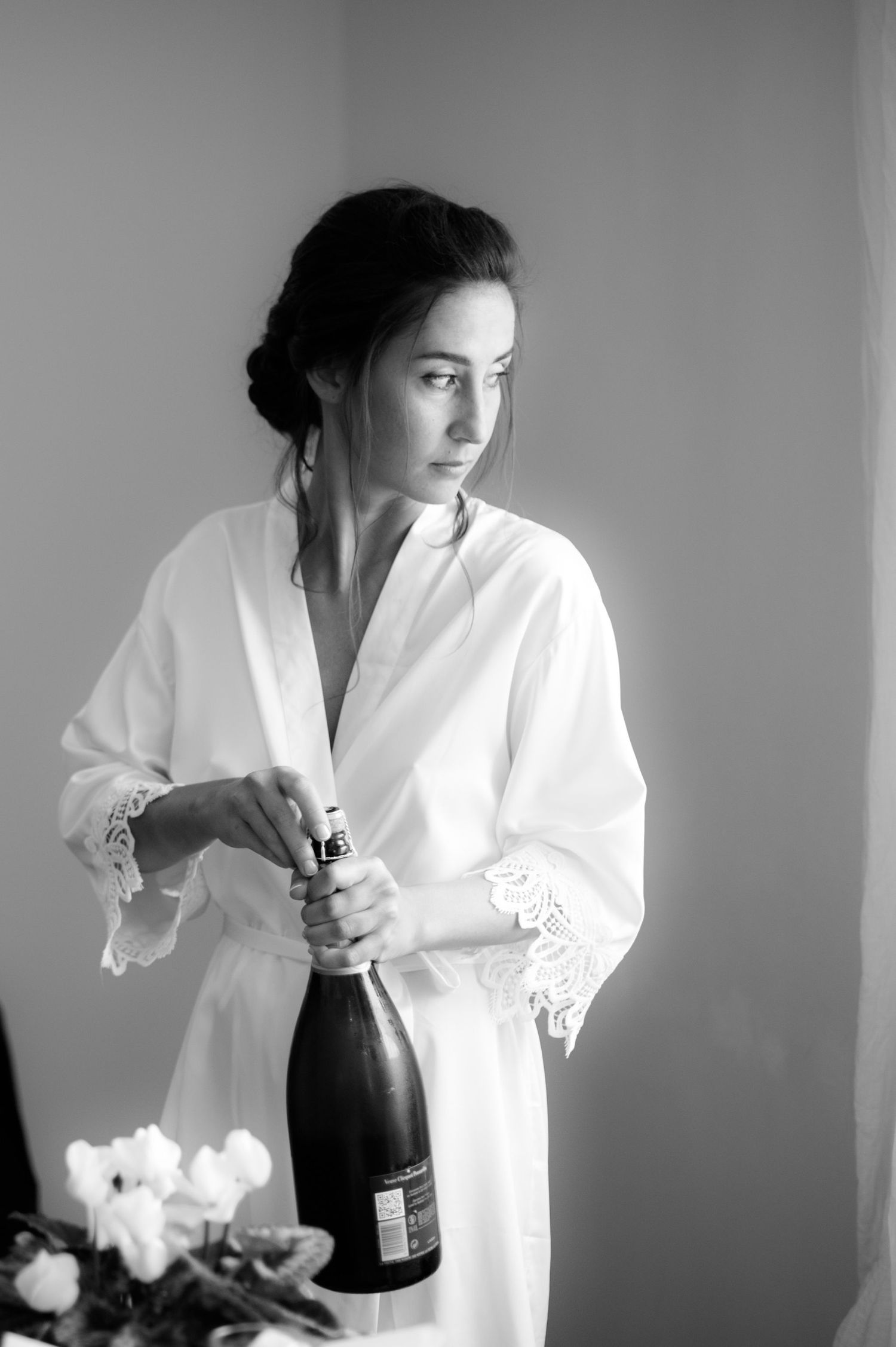 A person in white lace robe holds a champagne bottle in dramatic black and white lighting.