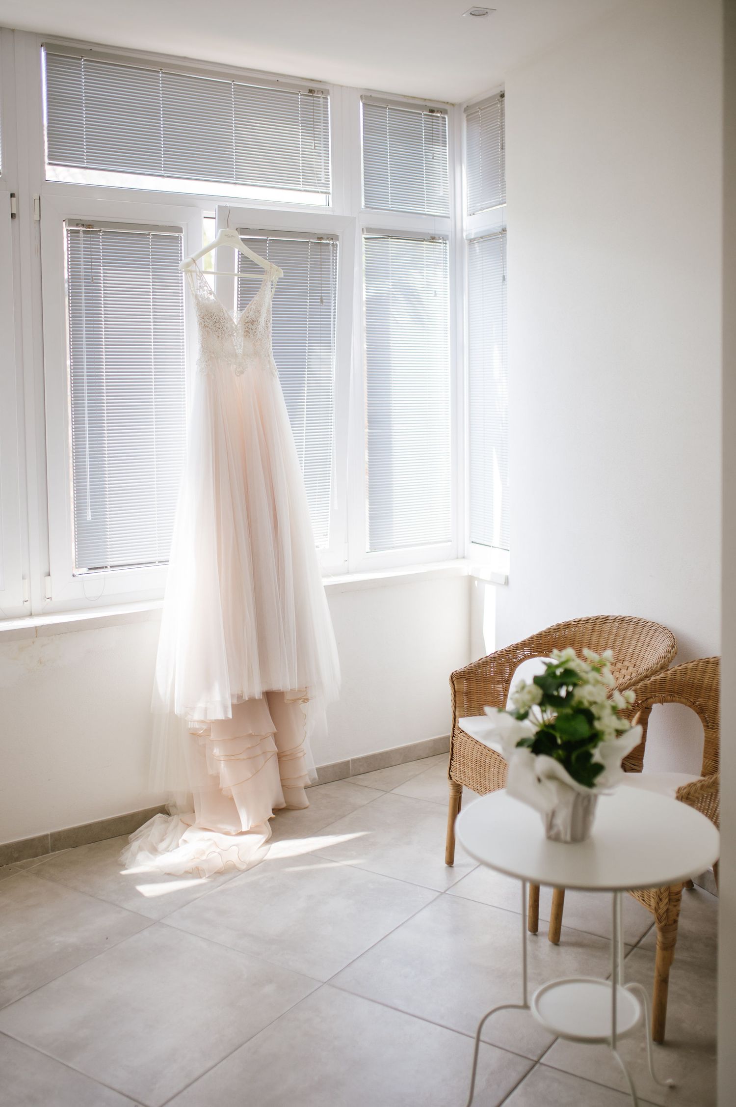 A wedding dress hangs by bright windows with a wicker chair and white floral bouquet.