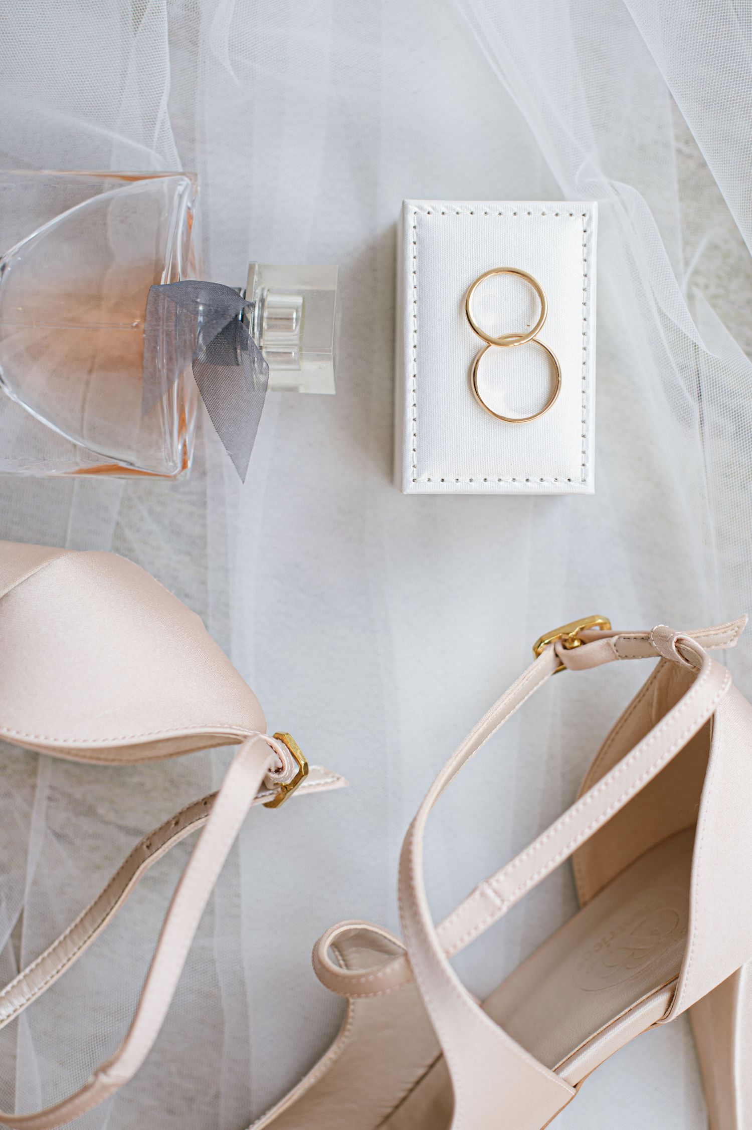 Wedding accessories including white wedding rings, perfume bottle and elegant beige heeled sandals on white fabric.