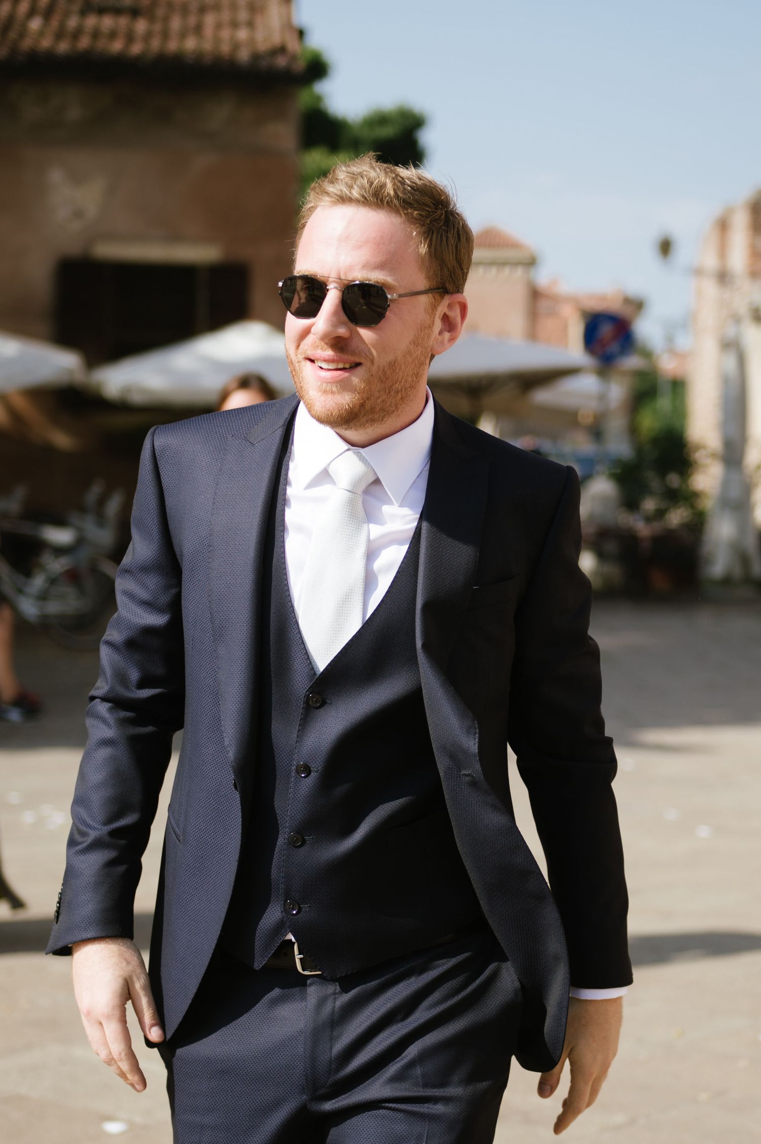 Stylish groom in navy suit and sunglasses walking through Italian streets.